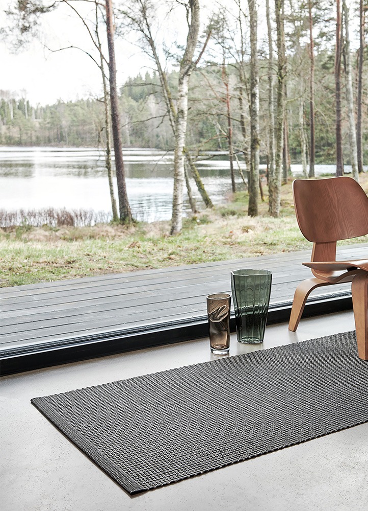 Un intérieur serein avec le tapis noir Pappelina Emm, placé près d'une grande fenêtre donnant sur un lac et des arbres. La scène comprend une chaise en bois et deux vases en verre sur le sol.